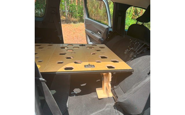 Innenraum eines Autos mit geöffneter Beifahrertür, rechteckige Holzplattform mit sechseckigen Öffnungen und einer Holzstütze auf den Rücksitzen, Bäume im Hintergrund