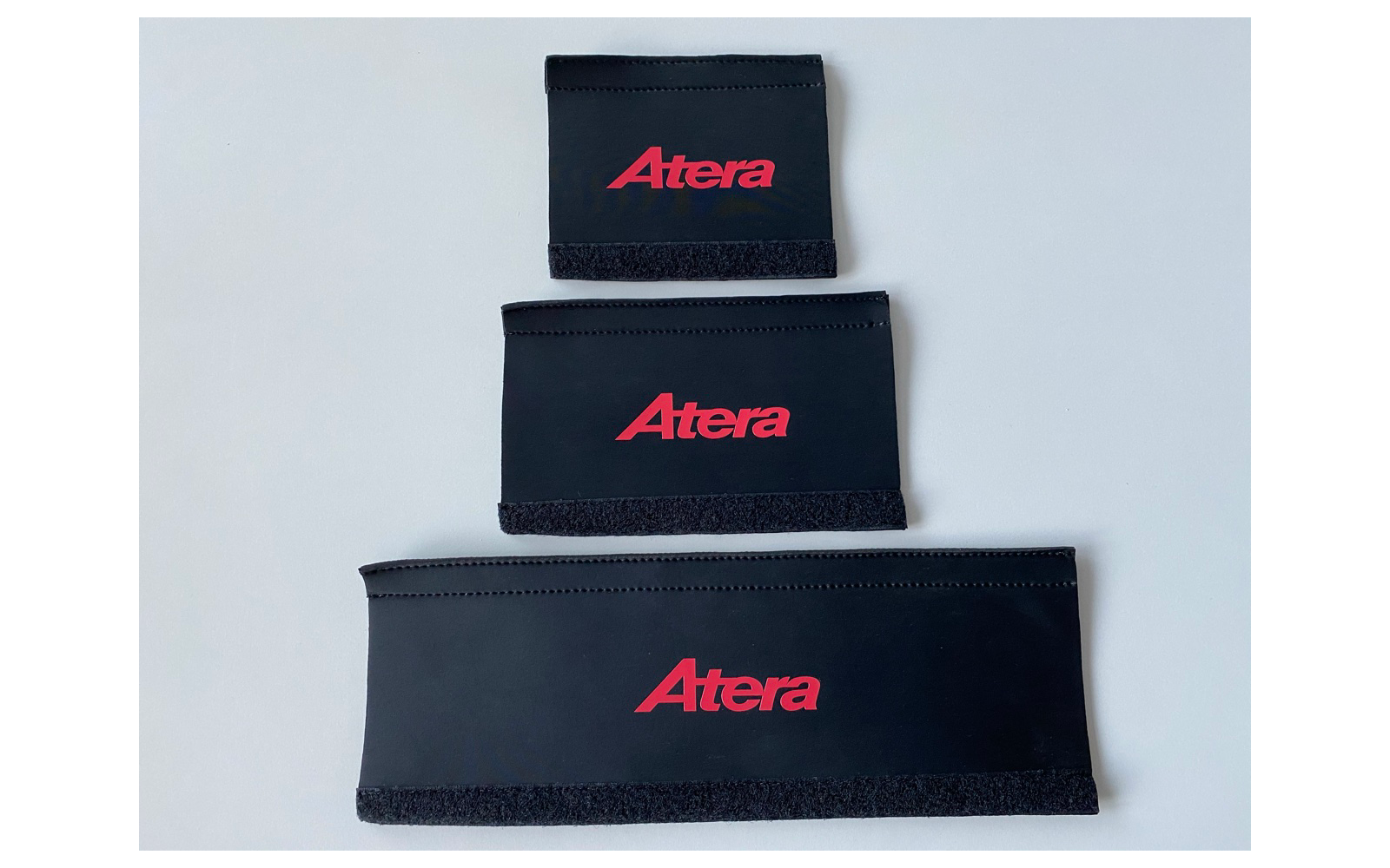 Drie zwarte rechthoekige stoffen banden met rood "Atera"-logo, verticaal gestapeld van klein naar groot op een lichte achtergrond