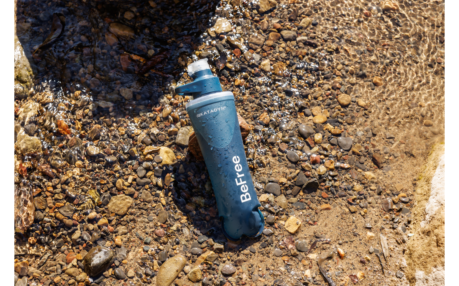 Blaue Flasche mit der Aufschrift 'BeFree' liegt auf kiesigem Ufer, teilweise im flachen Wasser