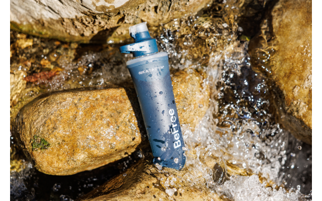 Blauer Behälter mit Schraubverschluss und Aufschrift 'BeFree' liegt zwischen nassen Steinen im fließenden Wasser