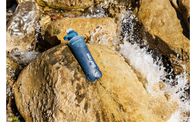 Blaue Trinkflasche mit weißer Aufschrift liegt auf einem nassen Felsen, Wasser fließt daneben