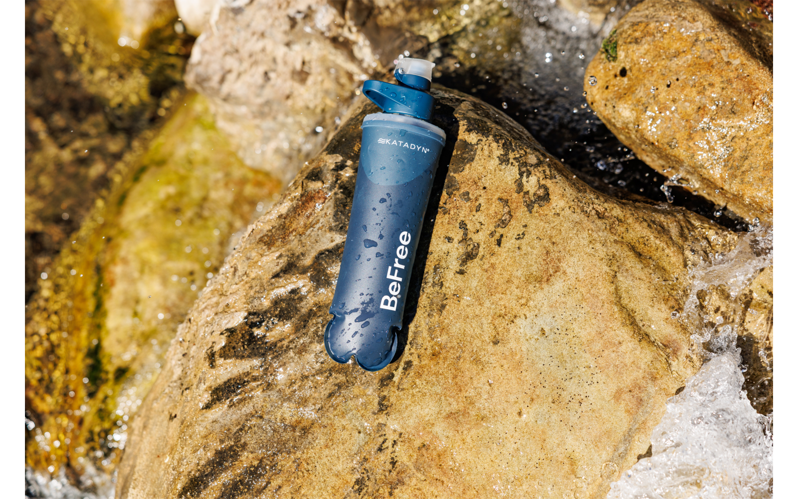 Blauer zylindrischer Gegenstand mit Aufschrift 'BeFree' liegt auf nassen Felsen, Wasser fließt daran vorbei