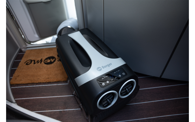 Klein zwart-wit apparaat met twee ronde ventilatieopeningen en een bedieningspaneel op een donkere houten vloer voor een vliegtuigdeur, naast een bruine deurmat met logo