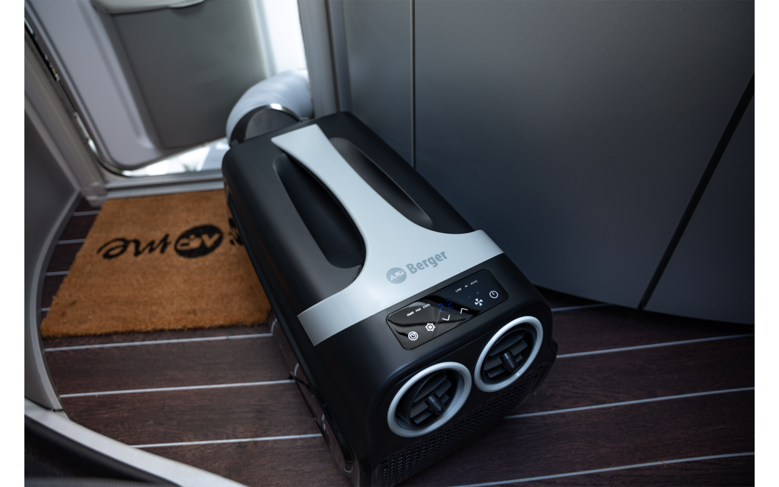 Klein zwart-wit apparaat met twee ronde ventilatieopeningen en een bedieningspaneel op een donkere houten vloer voor een vliegtuigdeur, naast een bruine deurmat met logo