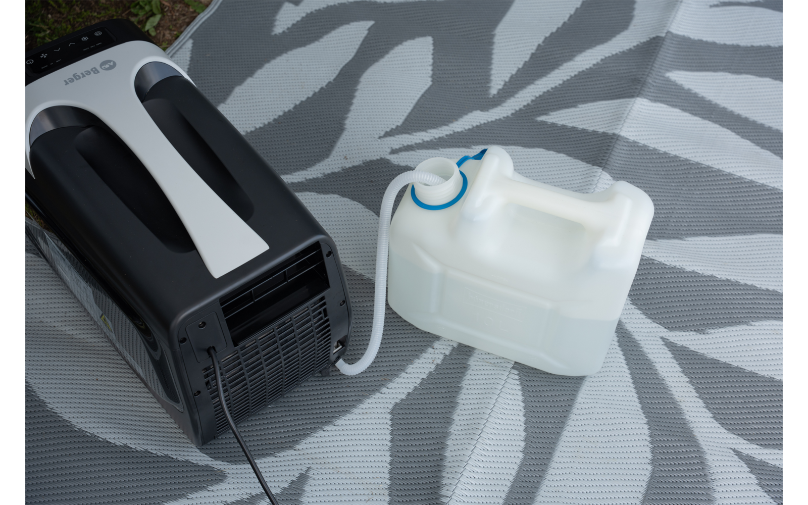 Mobiele airconditioner links met zwart-witte behuizing; een witte afvoerslang loopt naar een rechthoekige, doorschijnende witte watercontainer met blauwe schroefdop; apparaat en container staan op een grijze mat met een licht bladmotief