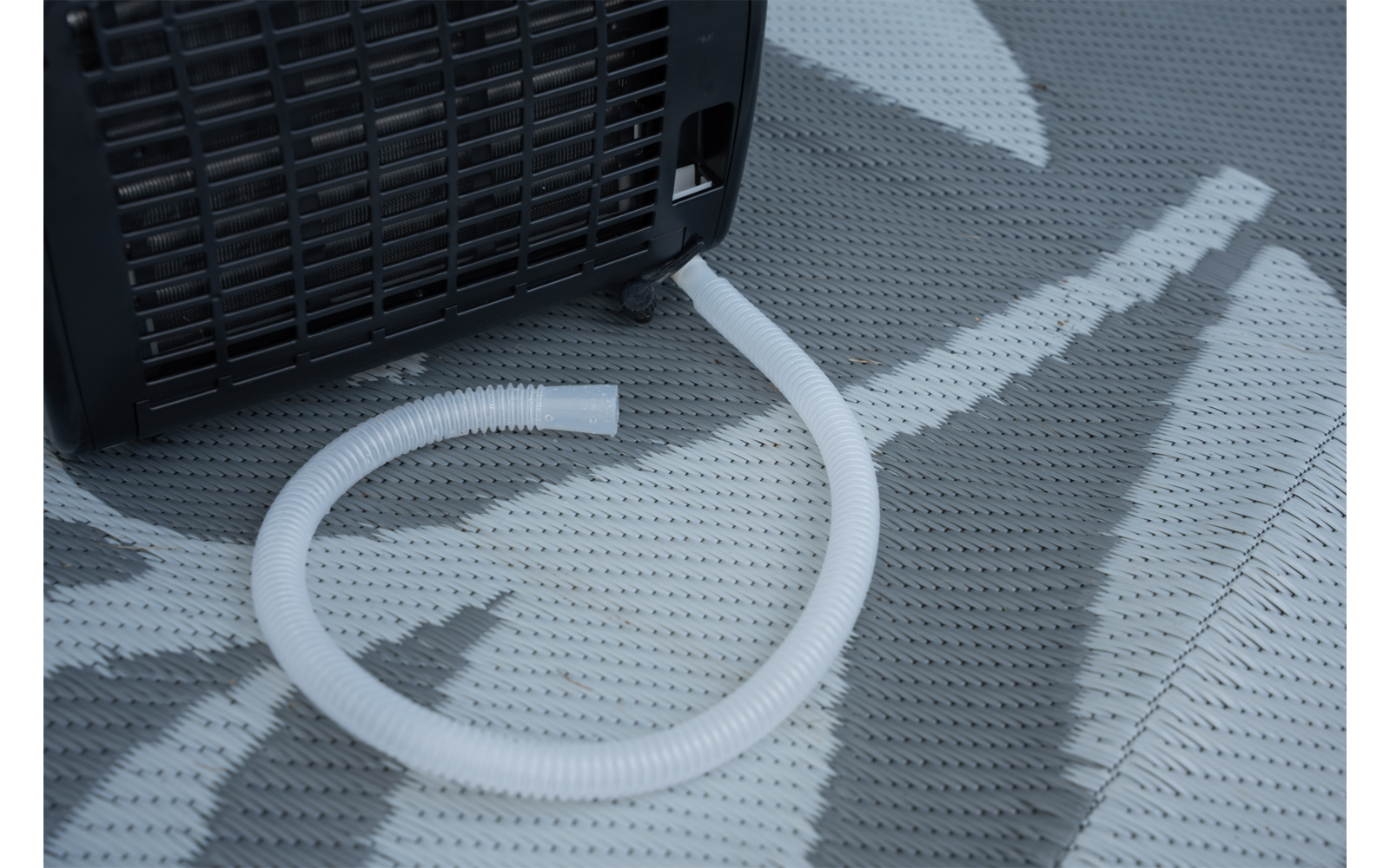 Zwart apparaat met ventilatierooster en witte geribbelde slang op een grijs vloerkleed met patroon