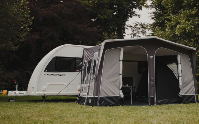 Wohnwagen mit angebautem grauem Vorzelt auf einer Wiese, umgeben von Bäumen
