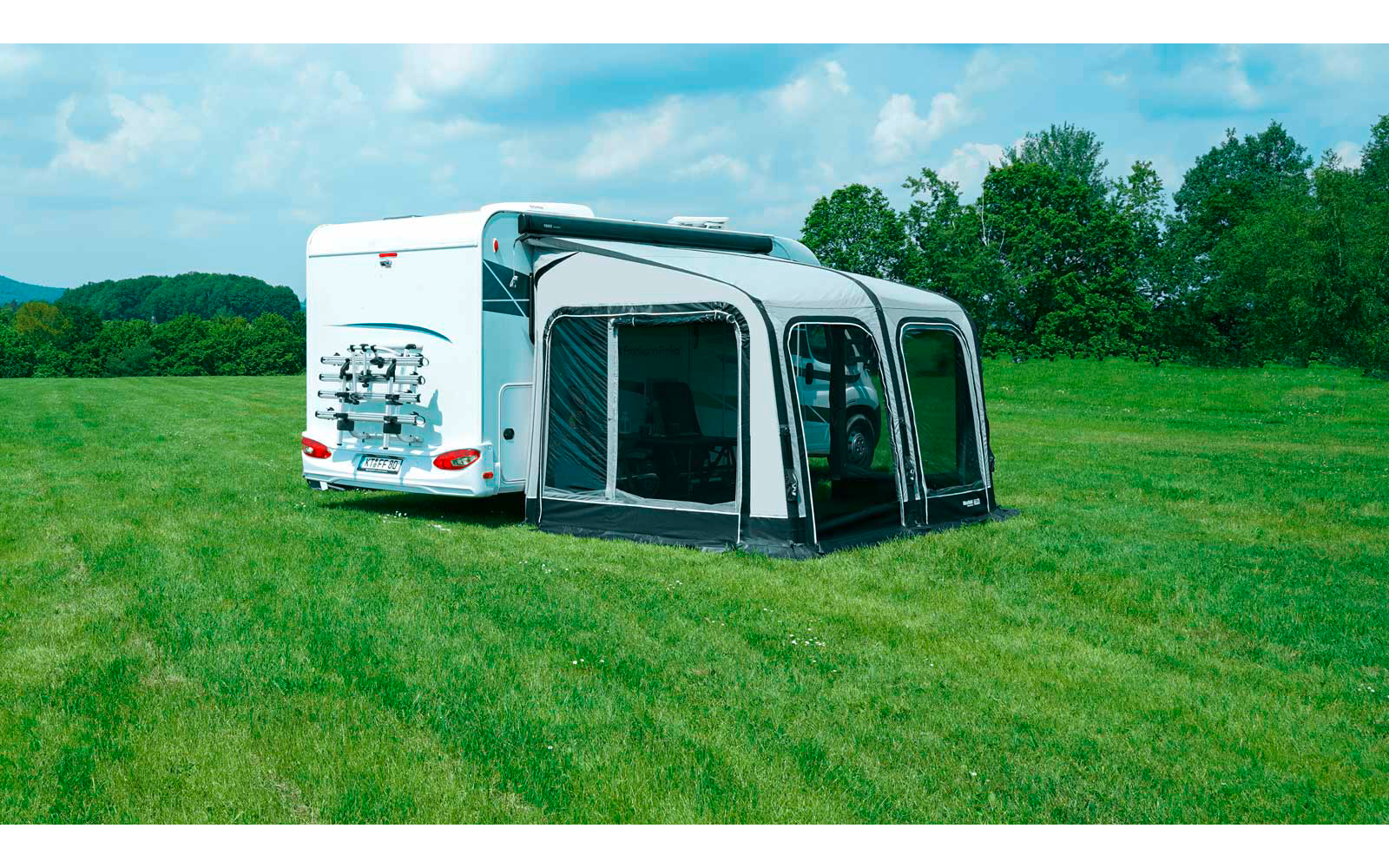 Weißes Wohnmobil mit grauem Vorzelt auf einer grünen Wiese, Bäume und wolkiger Himmel im Hintergrund