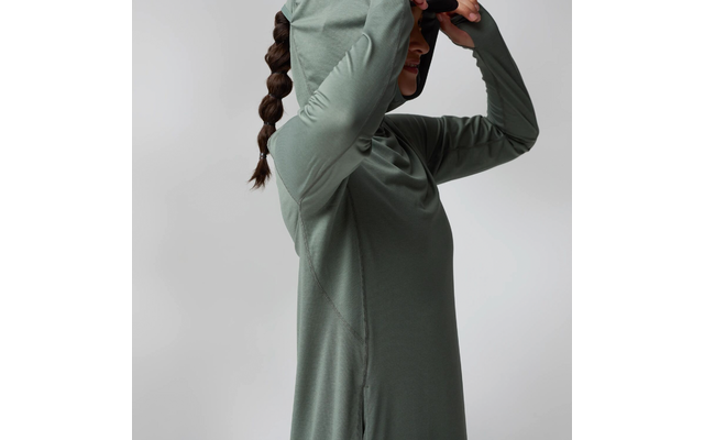 Personne avec tresse portée en queue de cheval portant un haut à capuche vert à manches longues, relève la capuche devant le visage ; vue de profil sur fond gris clair