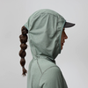 Personne de profil portant un sweat à capuche vert clair et une casquette noire à visière visible, longue tresse, fond uni clair