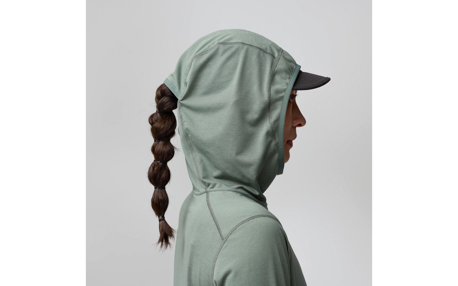 Personne de profil portant un sweat à capuche vert clair et une casquette noire à visière visible, longue tresse, fond uni clair