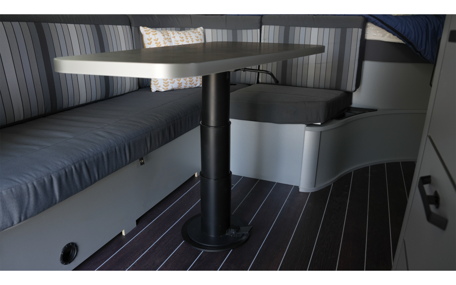 Bank mit blau-grau gestreiftem Polster, rechteckiger Tisch auf schwarzem Mittelfuß, weißes Kissen mit gelbem Muster, dunkler Boden mit hellen Streifen, Schranktüren mit Griffen, Matratzenkante mit Decke im Hintergrund