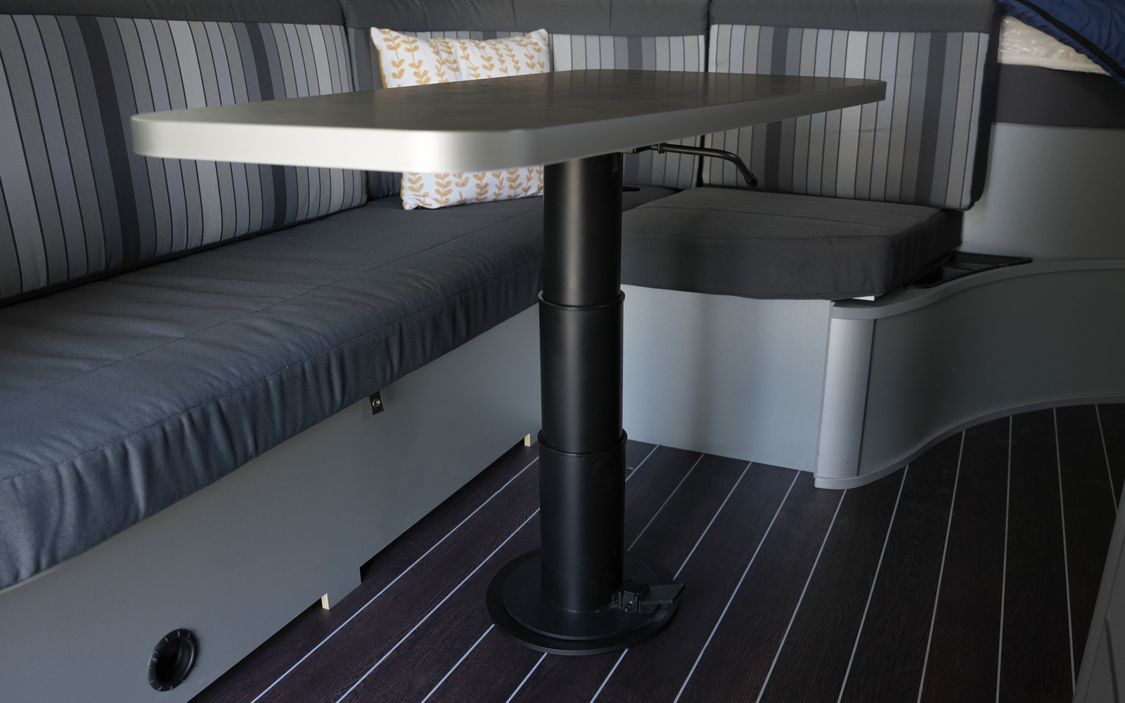 Banco con tapizado de rayas azul-gris, mesa rectangular sobre pedestal central negro, cojín blanco con motivo amarillo, suelo oscuro con rayas claras, puertas de armario con tiradores, borde del colchón con manta al fondo