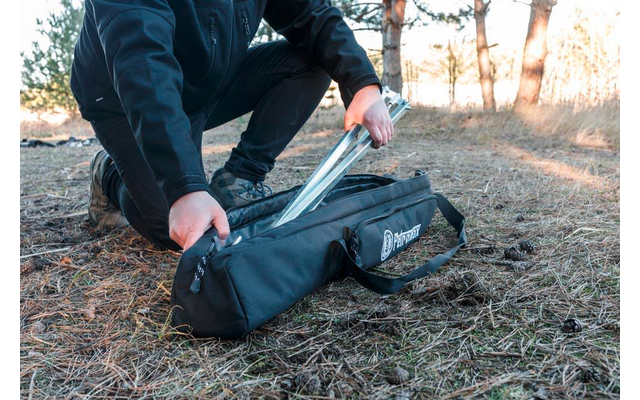 Person in schwarzer Jacke und dunkler Hose kniet auf trockenem Gras und hält zusammengesteckte Metallstangen über einer schwarzen Tragetasche; Bäume im Hintergrund