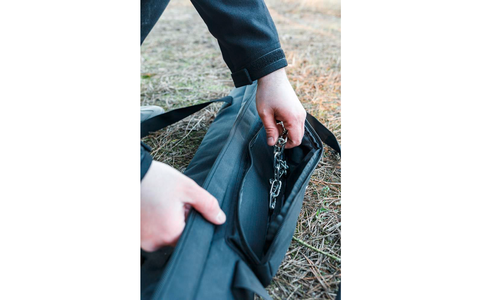 Zwei Hände öffnen eine schwarze Tasche auf Gras und halten eine Metallkette