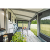 Vista del interior de una tienda anexa a una autocaravana: alfombra en el suelo, dos sillas de camping azules, mesa pequeña con bebidas, planta en maceta en primer plano, ventana lateral abierta con vista a un prado y árboles