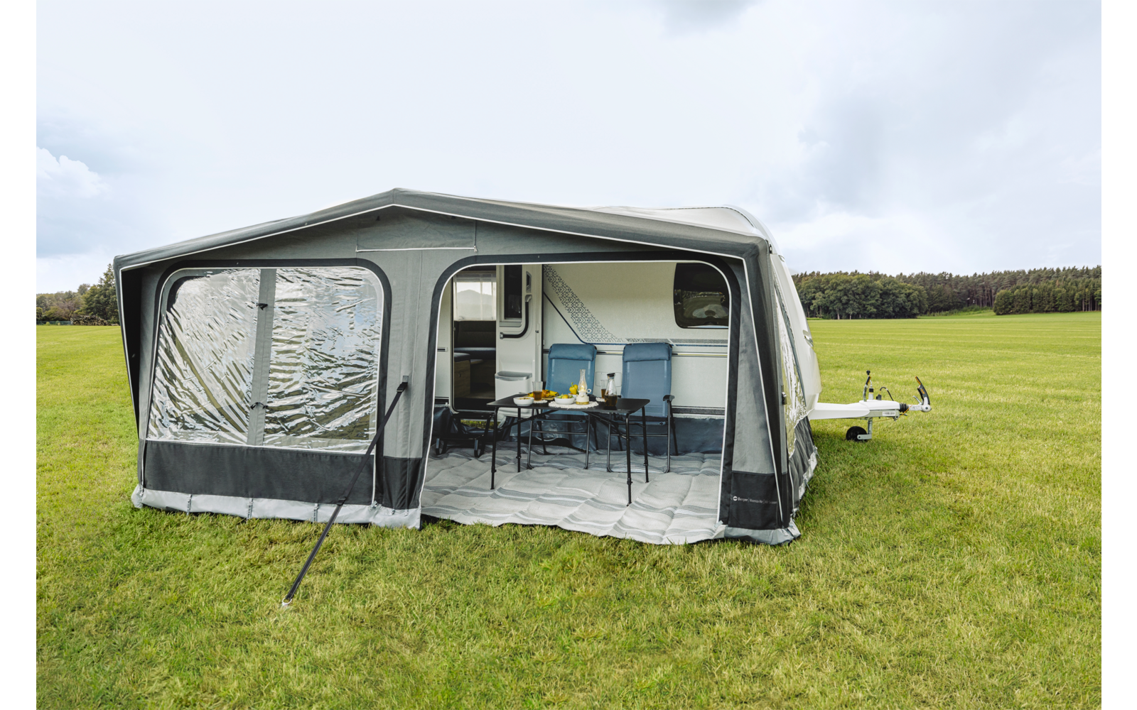 Toldo montado en una caravana con ventana de plástico transparente, entrada abierta, mesa con cuatro sillas y pequeña cocina; campo de hierba y cielo nublado al fondo