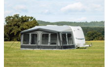 Witte caravan met aangebouwde grijze voortent op een grasveld, bomen en bewolkte lucht op de achtergrond