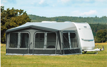 White caravan with gray awning on a grassy field, trees and forested hills in the background under a blue sky