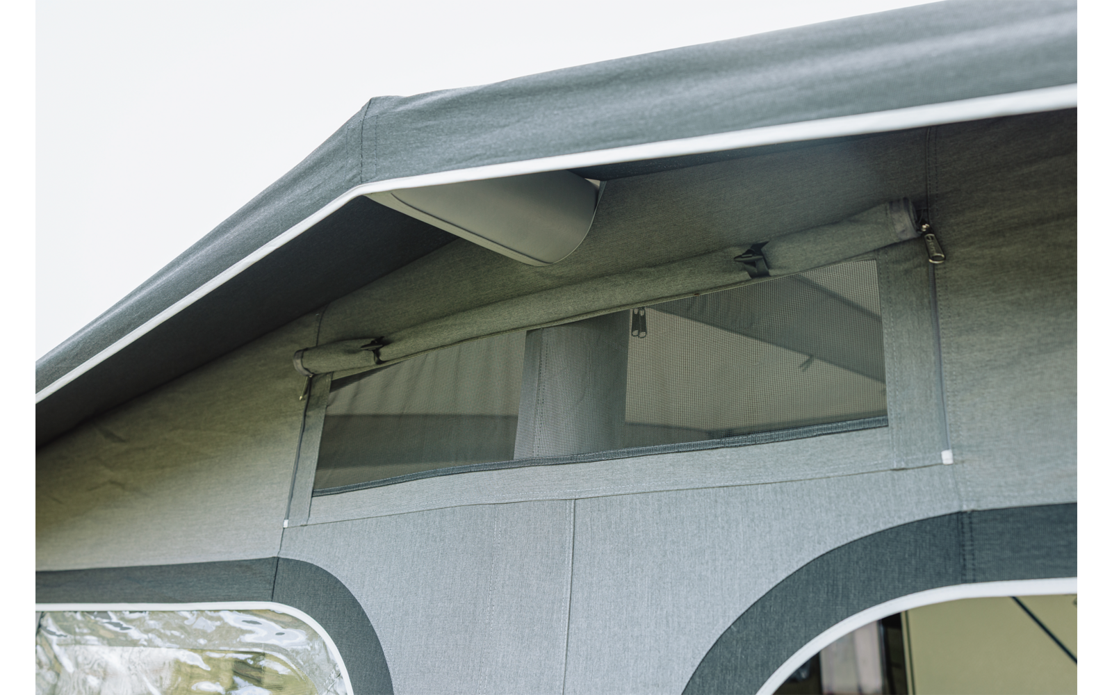 Estructura de tela gris con techo saliente, ventana rectangular transparente con capa de malla y cierres de cremallera, varillas metálicas visibles y dos paneles de tela semicirculares más oscuros debajo
