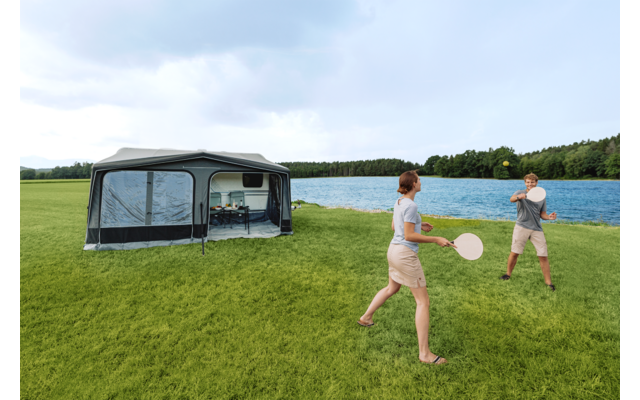 Dos personas con palas y una pelota juegan en un césped verde junto a un lago; a la izquierda una gran marquesina de camping gris con ventanas, al fondo árboles y cielo nublado