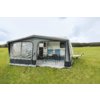 Toldo montado en una caravana con ventana de plástico transparente, entrada abierta, mesa con cuatro sillas y pequeña cocina; campo de hierba y cielo nublado al fondo