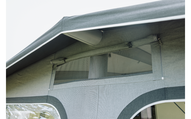 Estructura de tela gris con techo saliente, ventana rectangular transparente con capa de malla y cierres de cremallera, varillas metálicas visibles y dos paneles de tela semicirculares más oscuros debajo