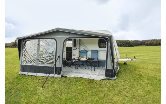 Toldo montado en una caravana con ventana de plástico transparente, entrada abierta, mesa con cuatro sillas y pequeña cocina; campo de hierba y cielo nublado al fondo