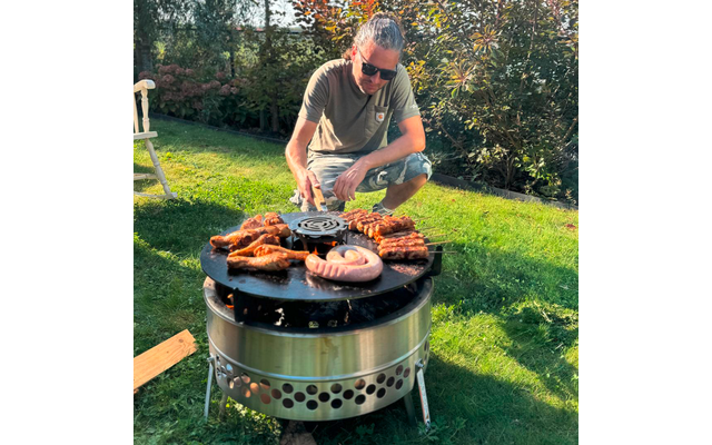 Person mit Sonnenbrille und grünem T-Shirt hockt neben rundem Metallgrill auf Rasen; auf der Grillplatte liegen Würste, Hähnchenteile, Rippchen und Spieße; Hecke und Gartenstuhl im Hintergrund