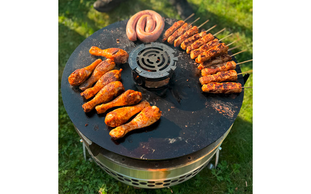 Runde Grillplatte mit mehreren Hähnchenkeulen unten links, einer spiraligen Bratwurst oben, Fleischspießen rechts und einem zentralen Lüftungsgitter, auf Gras