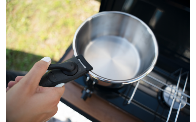 Eine Hand hält einen schwarzen abnehmbaren Griff an einen silbernen Edelstahltopf, der auf einem tragbaren Gaskocher mit offenem Brenner steht; im Hintergrund Gras