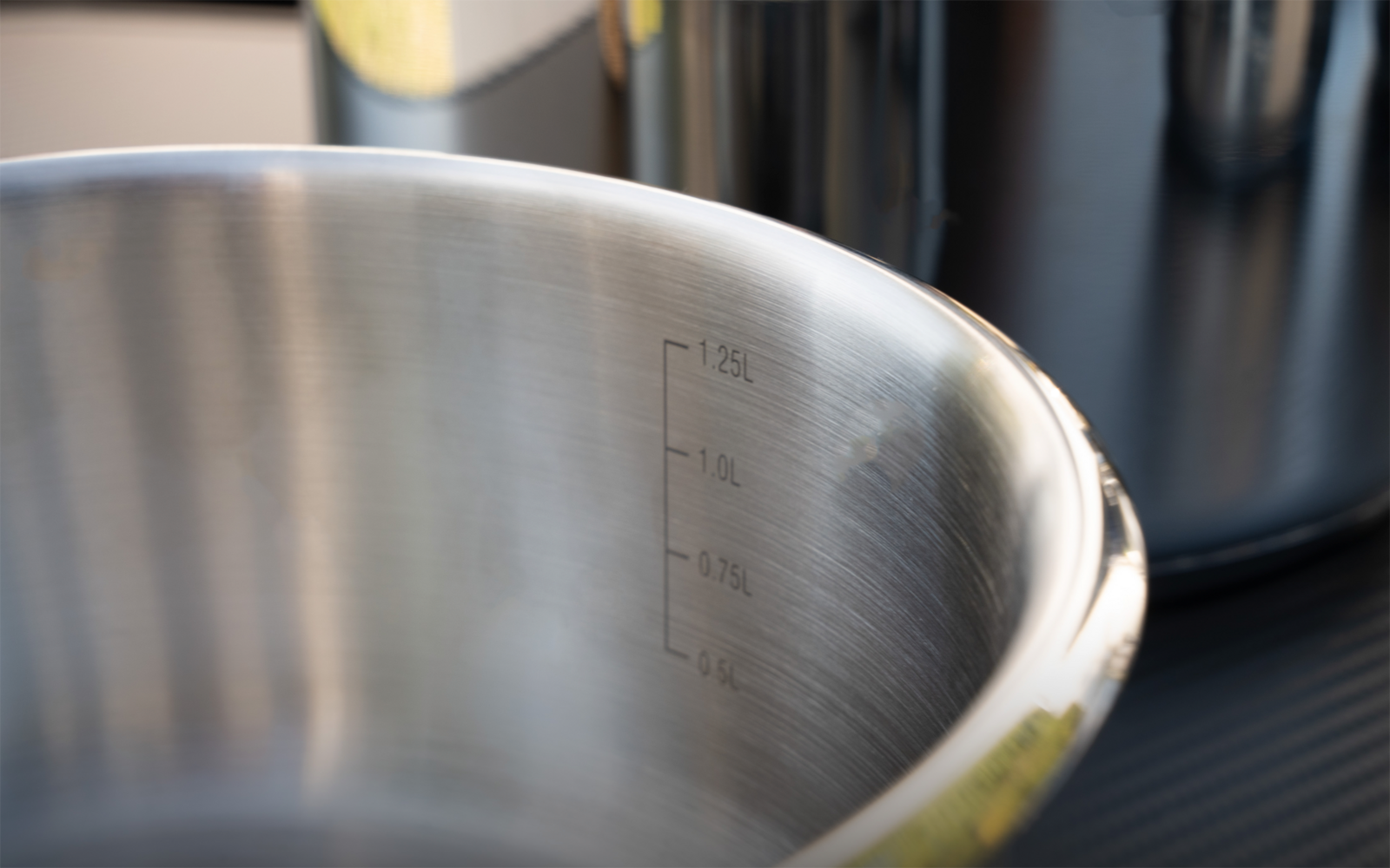 Edelstahlschüssel seitlich, innen sichtbare Messskala mit Zahlen, glänzender Rand; im Hintergrund ein Edelstahltopf auf dunkler Oberfläche