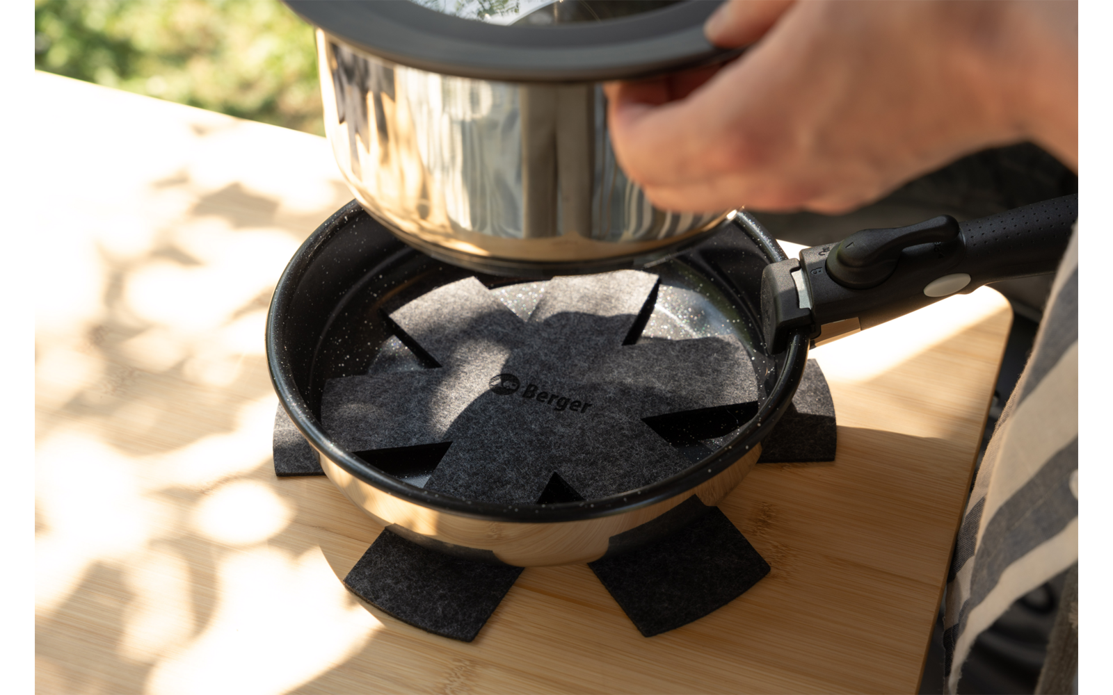 Two hands hold a metal pot over a small black frying pan; a star-shaped felt pad sits inside the pan; several felt squares are placed around the pan; pan on a wooden surface, blurred green background
