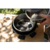 Hand pours water from a metal kettle into a round black bowl with a cross-shaped insert on a wooden board; blurred garden background