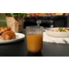 Glas mit Orangensaft auf dunklem Tisch; im Hintergrund links ein Teller mit Croissants, ein geflochtener Korb mit Pflanze, rechts eine Schüssel mit Salat auf einer Spitzenunterlage