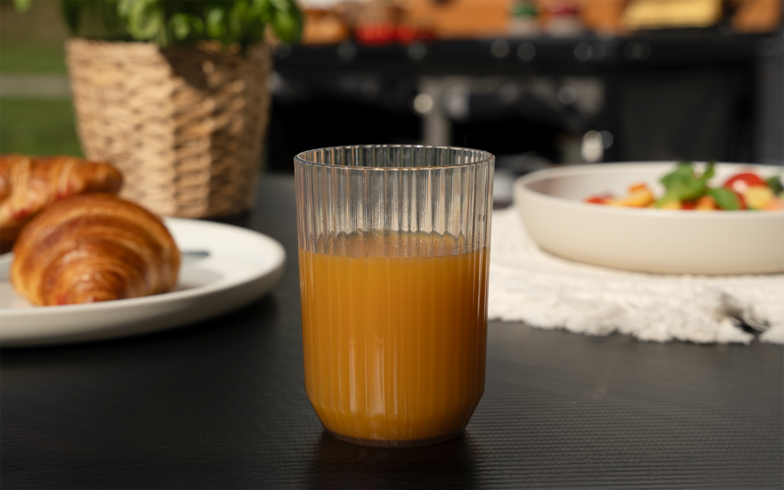 Glas mit Orangensaft auf dunklem Tisch; im Hintergrund links ein Teller mit Croissants, ein geflochtener Korb mit Pflanze, rechts eine Schüssel mit Salat auf einer Spitzenunterlage