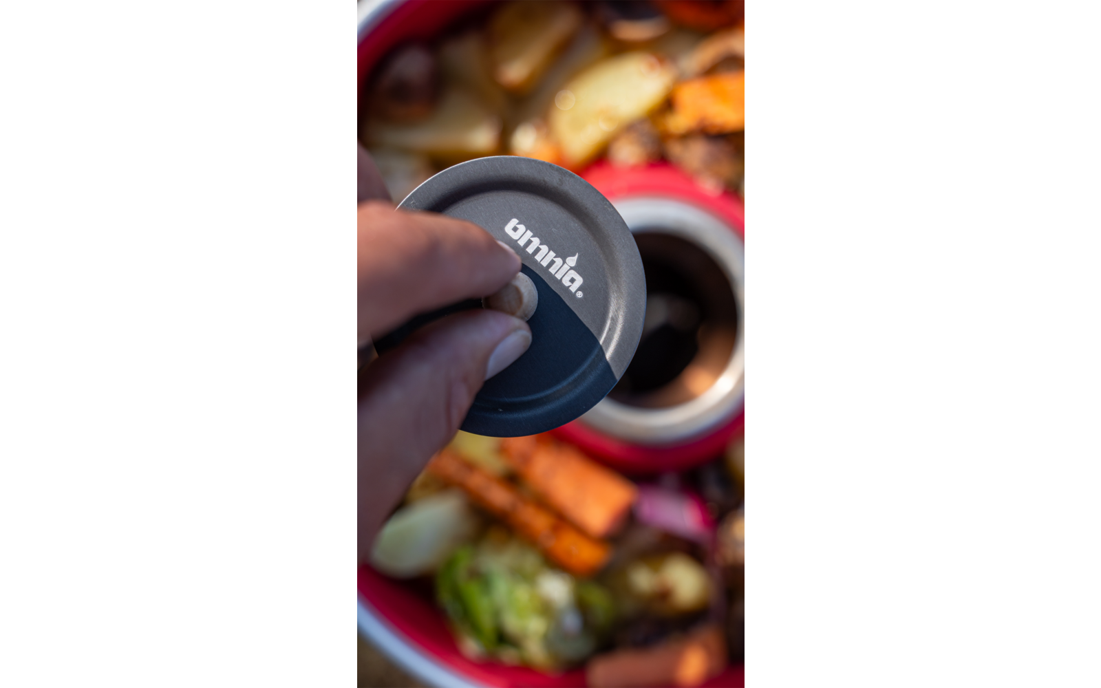 Main tenant un couvercle gris à texte blanc au-dessus d'un récipient rouge contenant des légumes