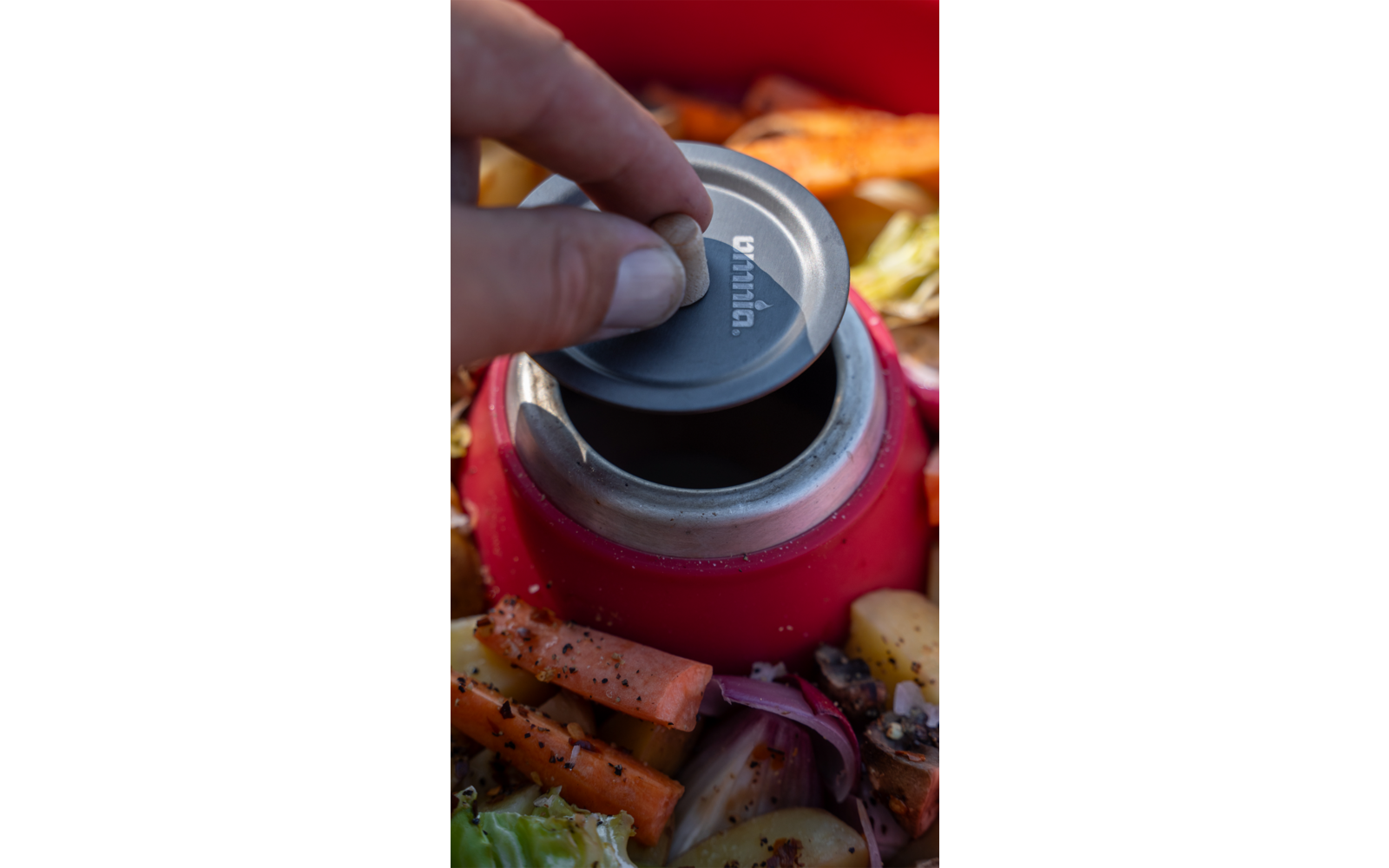 Des doigts tiennent un couvercle métallique rond au-dessus de l'ouverture d'un récipient rouge, entouré de morceaux de carotte, de feuilles de chou et de tranches d'oignon rouge