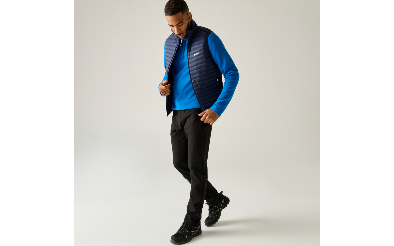 Persoon met blauwe trui, donkeblauwe doorgestikte bodywarmer, zwarte broek en zwarte wandelschoenen, staand voor een lichte achtergrond, kijkt omlaag en raakt de bodywarmer aan