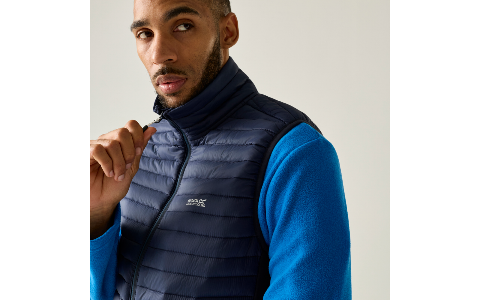 Persoon draagt donkerblau gewatteerde bodywarmer over een blauw fleece, houdt de rits bij de kin vast, kijkt opzij, egale lichte achtergrond