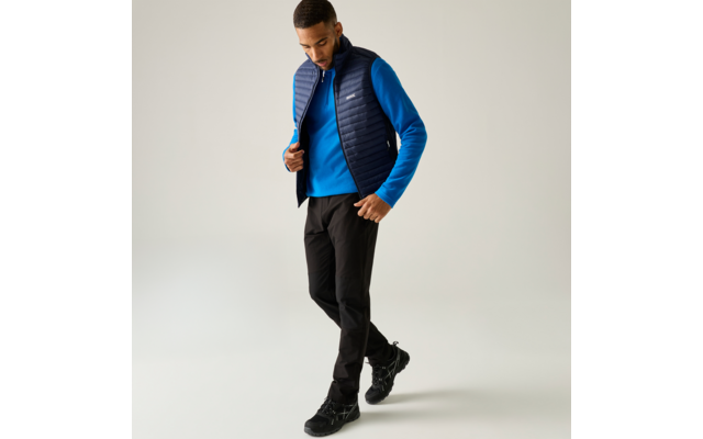 Persoon met blauwe trui, donkeblauwe doorgestikte bodywarmer, zwarte broek en zwarte wandelschoenen, staand voor een lichte achtergrond, kijkt omlaag en raakt de bodywarmer aan