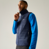Man met blauw shirt met lange mouwen en donkerblauw gewatteerd vest, houdt rits bij de kraag vast, kijkt omhoog tegen een lichte achtergrond