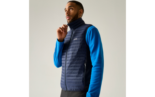 Man met blauw shirt met lange mouwen en donkerblauw gewatteerd vest, houdt rits bij de kraag vast, kijkt omhoog tegen een lichte achtergrond