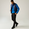 Persoon met blauwe trui, donkeblauwe doorgestikte bodywarmer, zwarte broek en zwarte wandelschoenen, staand voor een lichte achtergrond, kijkt omlaag en raakt de bodywarmer aan