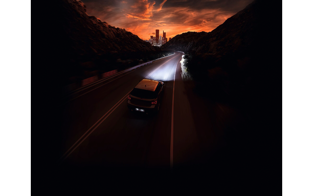 Achteraanzicht van een SUV op een tweebaansweg tussen rotsachtige hellingen, tegemoetkomende koplampen, verlichte stadsachtergrond en oranje wolken aan de hemel