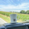 Weiße Tasse mit Kompassrose auf dem Armaturenbrett, Straße, Maisfeld und blauer Himmel mit Wolken im Hintergrund