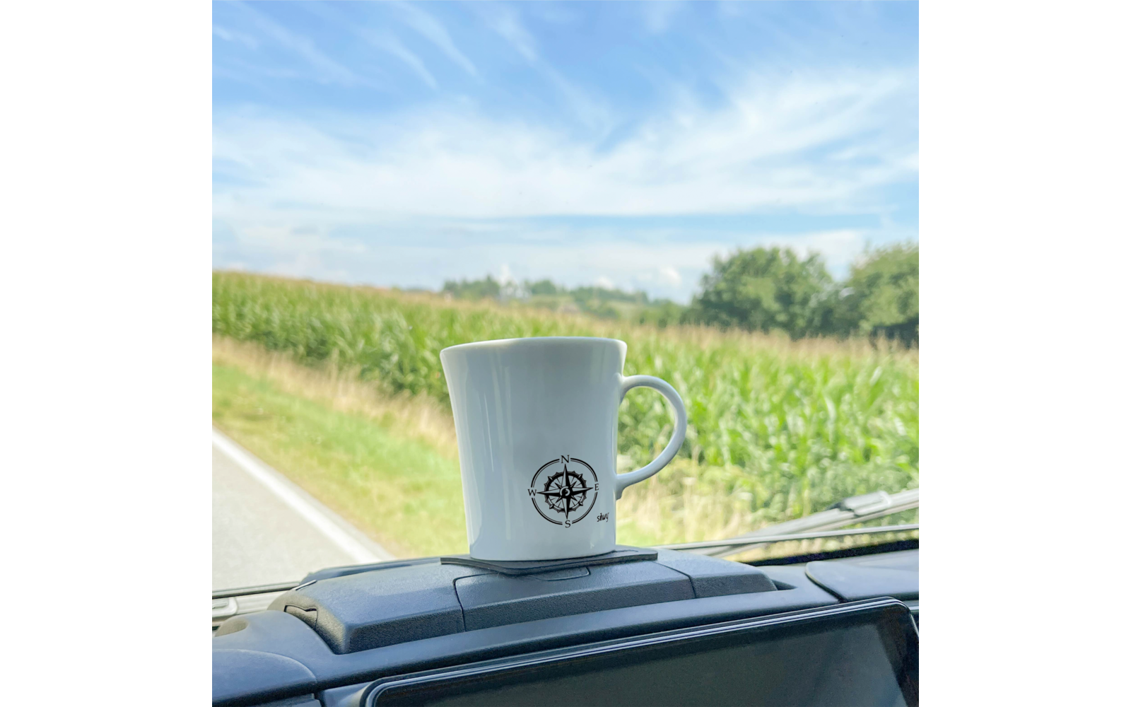 Weiße Tasse mit Kompassrose auf dem Armaturenbrett, Straße, Maisfeld und blauer Himmel mit Wolken im Hintergrund
