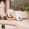 Zwei weiße Tassen mit Kompass-Logo auf Küchenarbeitsplatte; eine Tasse auf Holzhalter, daneben kleine Pflanze, links Handtuch an Haken, im Hintergrund nasses Fenster mit Bäumen