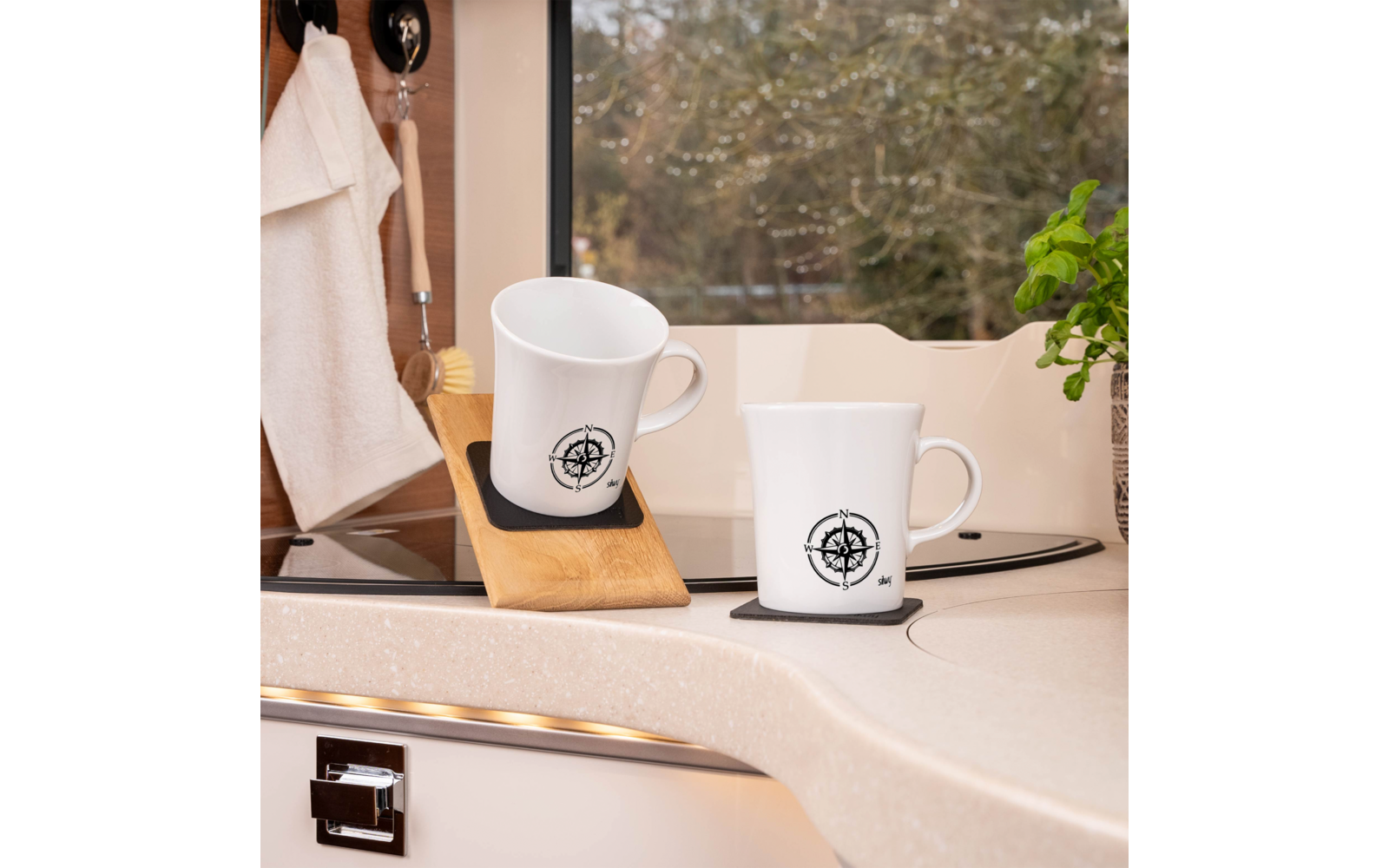 Zwei weiße Tassen mit Kompass-Logo auf Küchenarbeitsplatte; eine Tasse auf Holzhalter, daneben kleine Pflanze, links Handtuch an Haken, im Hintergrund nasses Fenster mit Bäumen