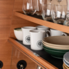 Holzschrankfach mit oben stielosen Gläsern, weißen Tassen mit schwarzem Logo und gestapelten grünen und beigen Schalen sowie Tellern